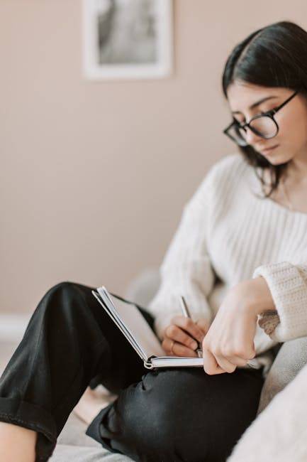 顯化感恩日記的進階技巧 Attentive female wearing eyeglasses and casual outfit sitting barefoot with crossed legs on comfortable couch in modern flat and taking notes in notebook with pen
